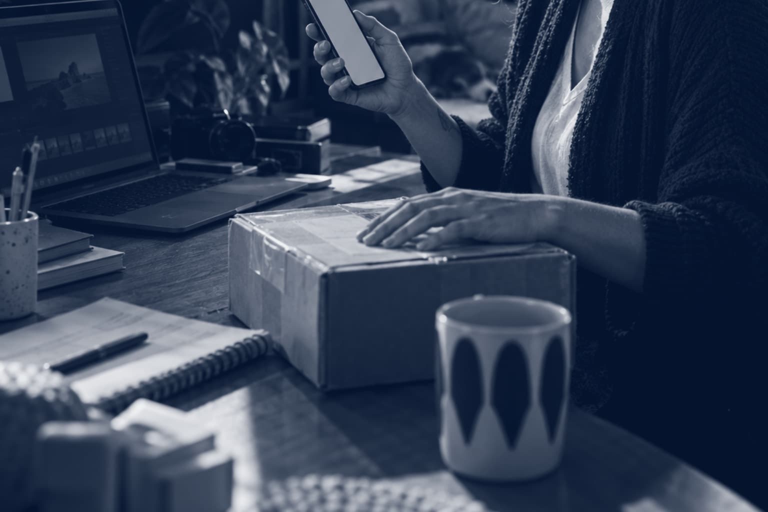 Mulher na mesa de casa com o celular numa mão e a outra sobre uma caixa de encomenda, ao lado do notebook e de uma caneca, em tons de azul