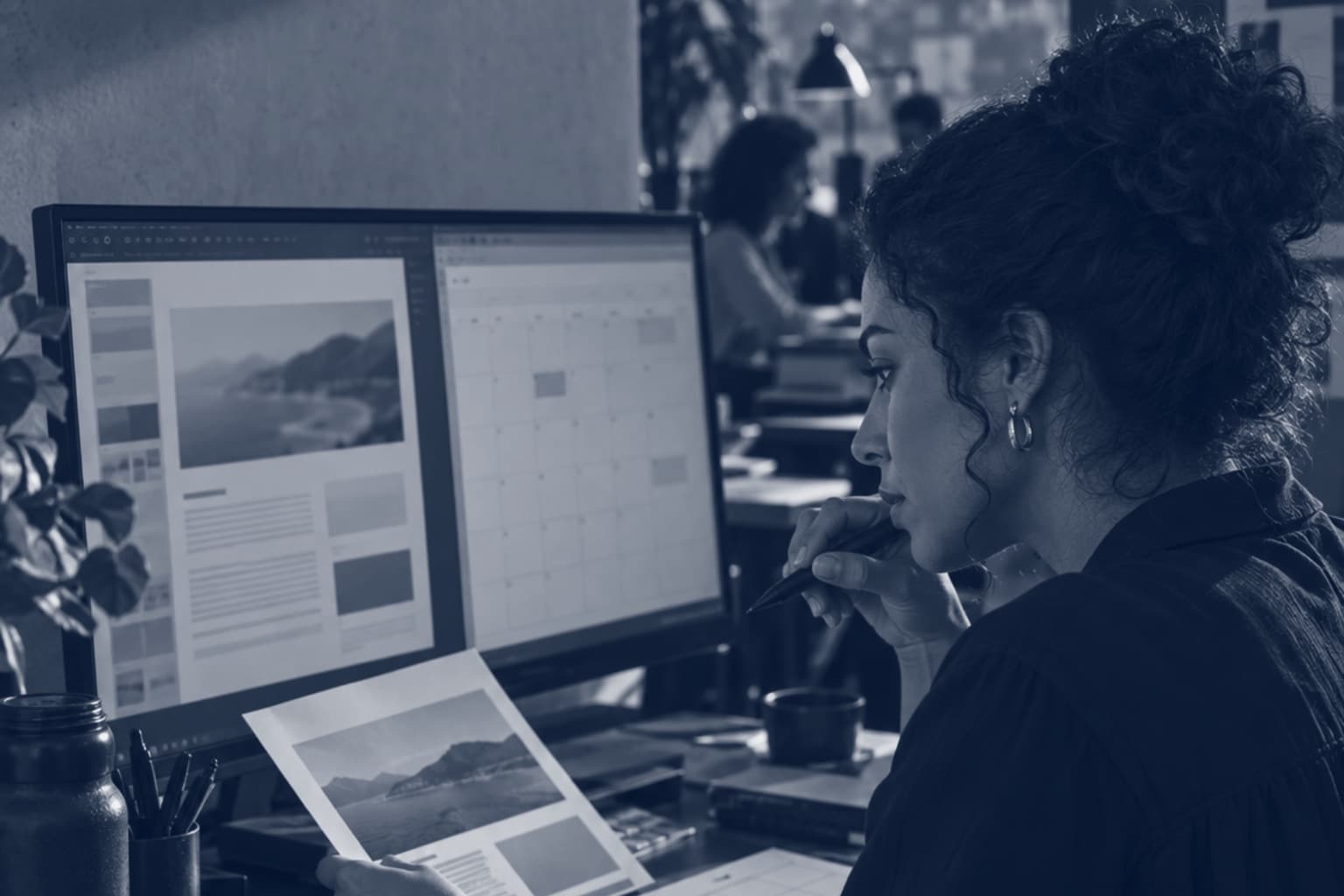 Designer de perfil diante do monitor, com a caneta no queixo e uma prova impressa na mão, conferindo o trabalho, em tons de azul